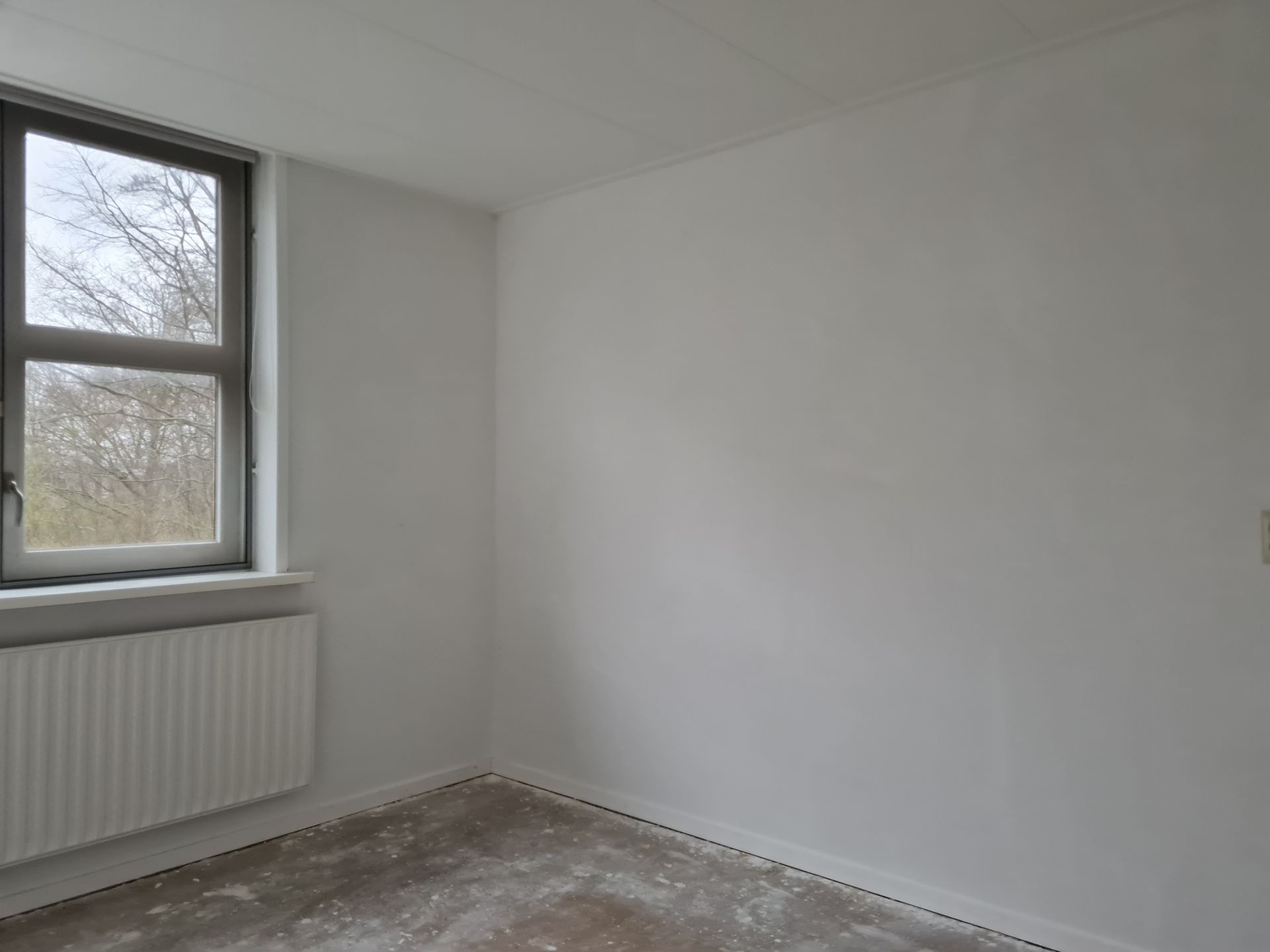 Een lege, witte kamer met een raam en radiator, een kale vloer en effen muren.