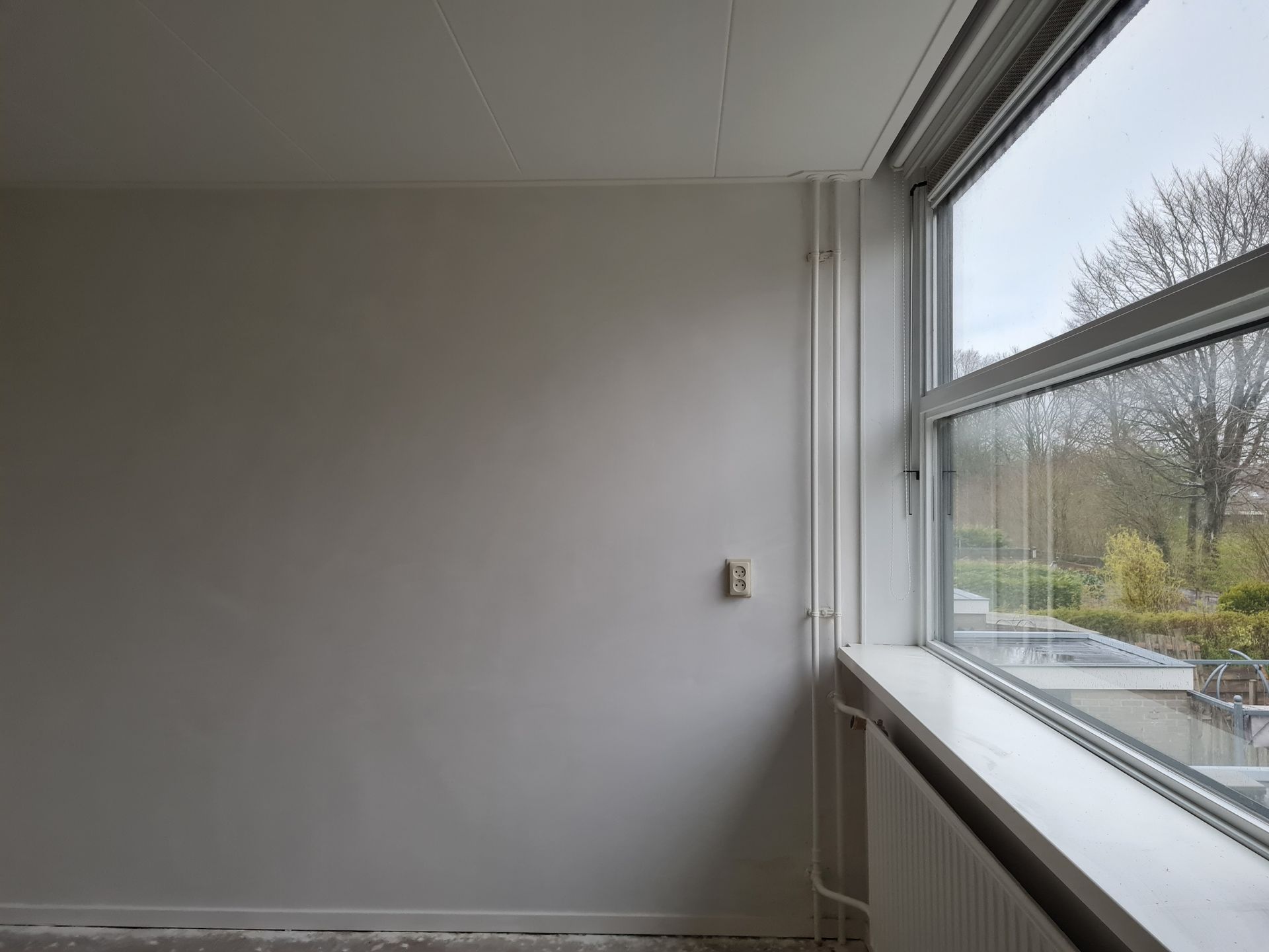Een lege kamer met grijze muren en een groot raam met uitzicht op bomen en geparkeerde auto's.