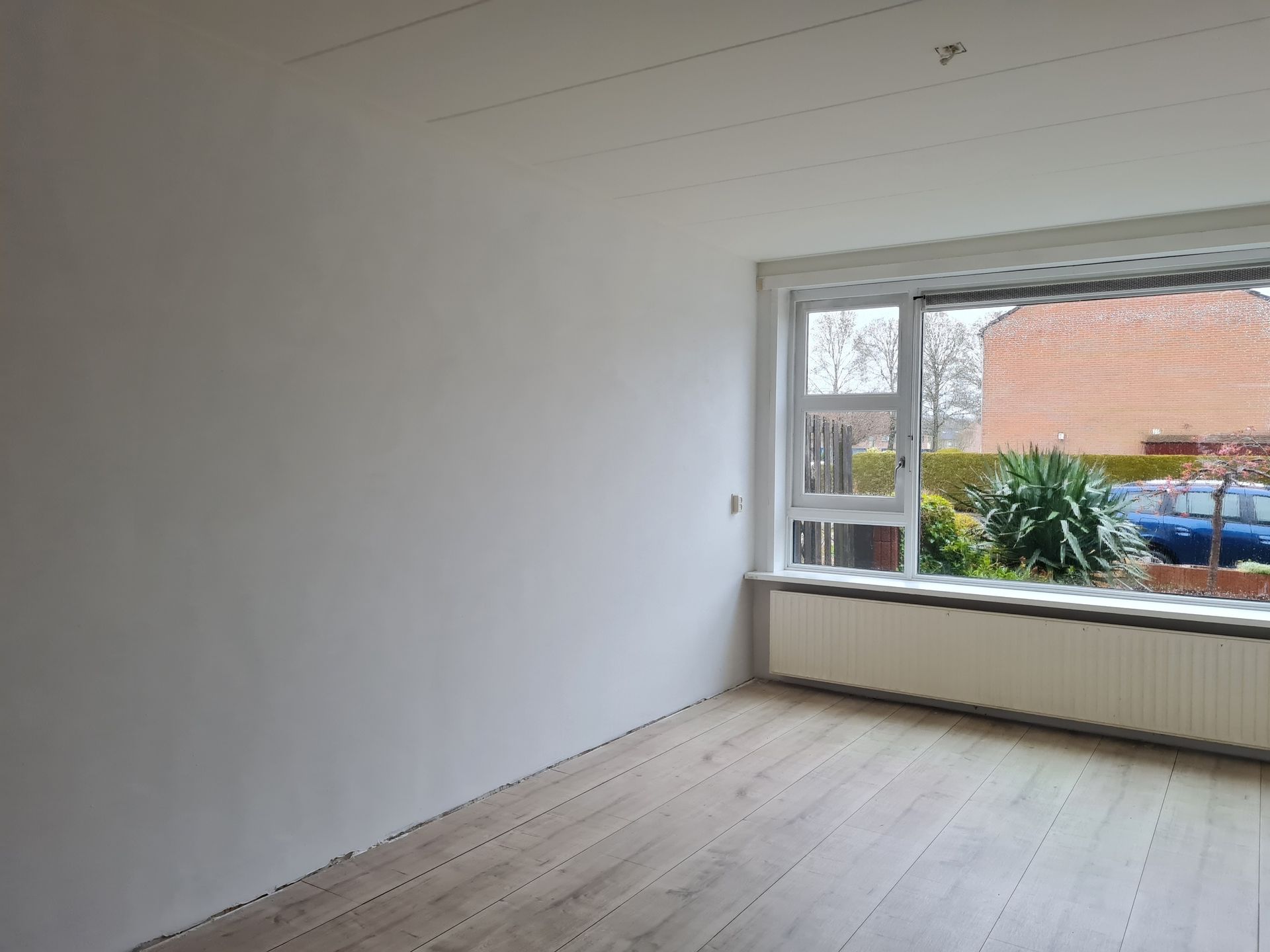 Lege witte kamer met houten vloer en een groot raam met uitzicht op groen en een rood dak.