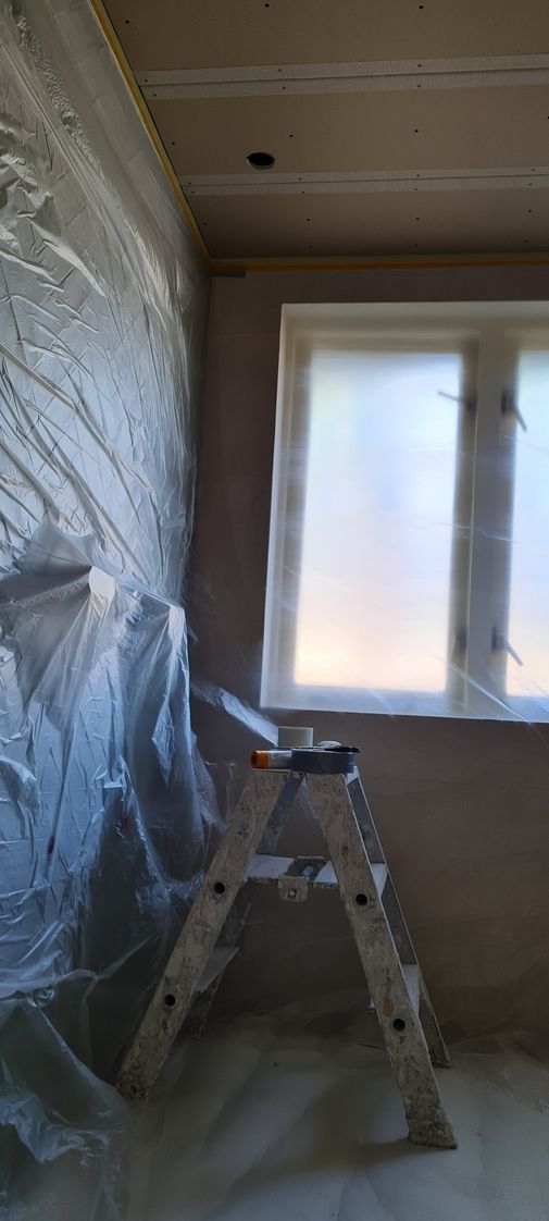 Renovatiekamer met met plastic beklede wand, trapladder en licht raam.