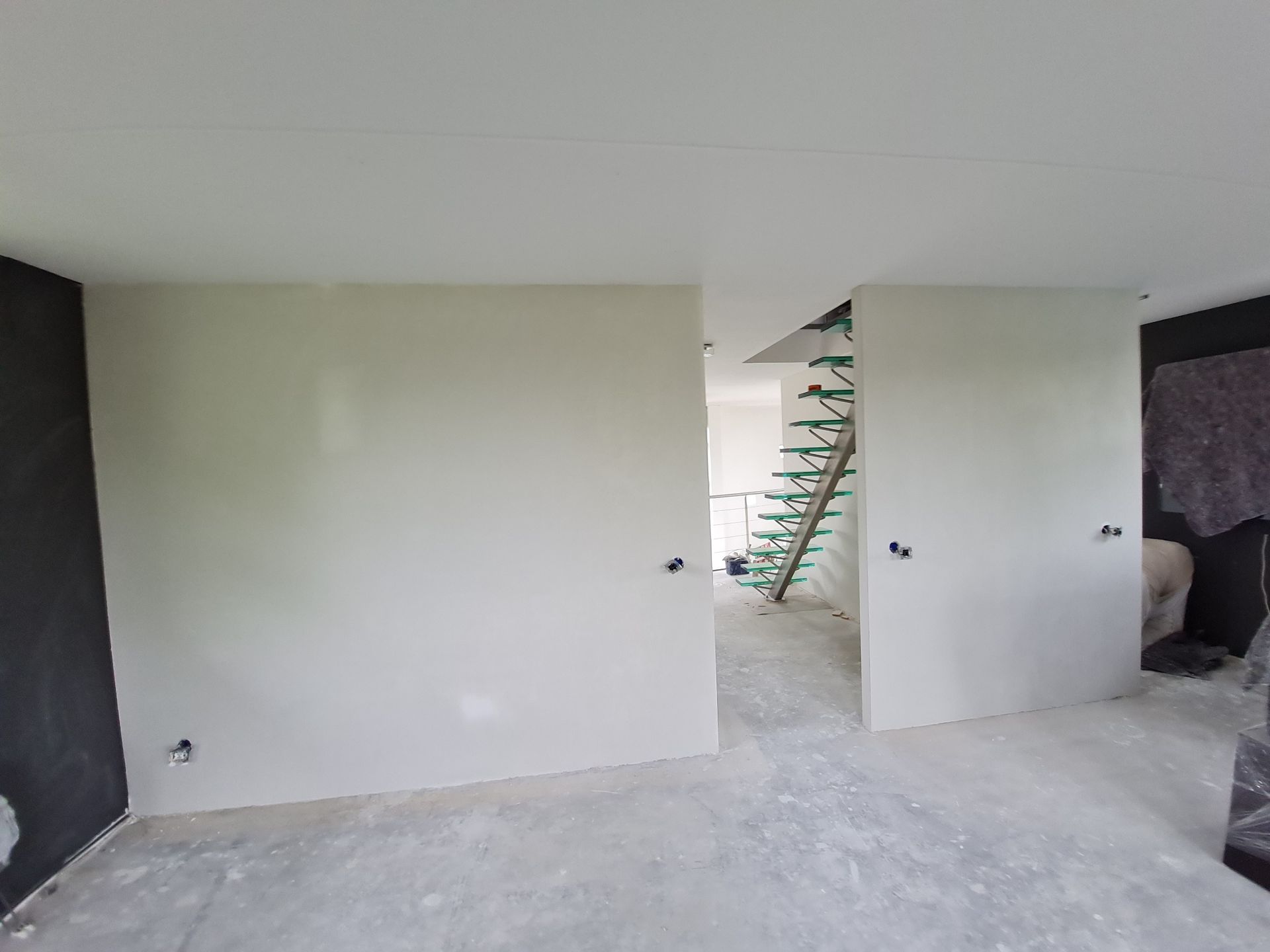 Een lege, onafgewerkte kamer met witte muren, een betonnen vloer en een trap die door een deuropening zichtbaar is.