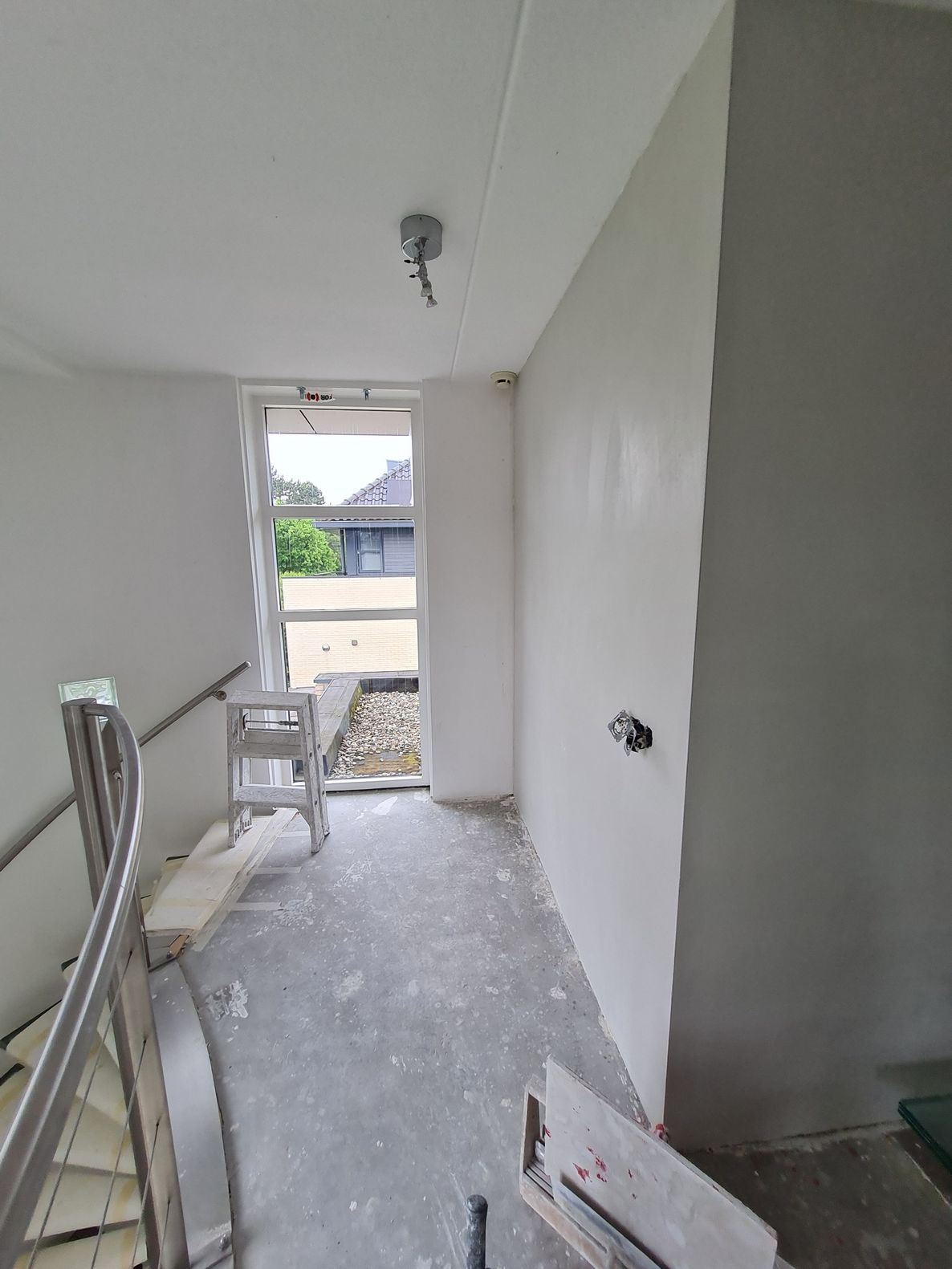 Smalle, onafgewerkte gang met witte muren, een betonnen vloer en een raam aan het uiteinde.