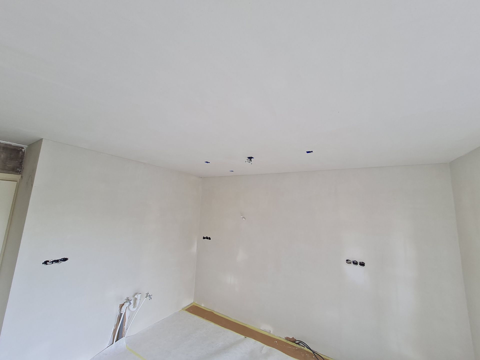 Lege witte kamer met onafgewerkte muren, plafondlampen en zichtbare leidingen vlak bij de vloer.