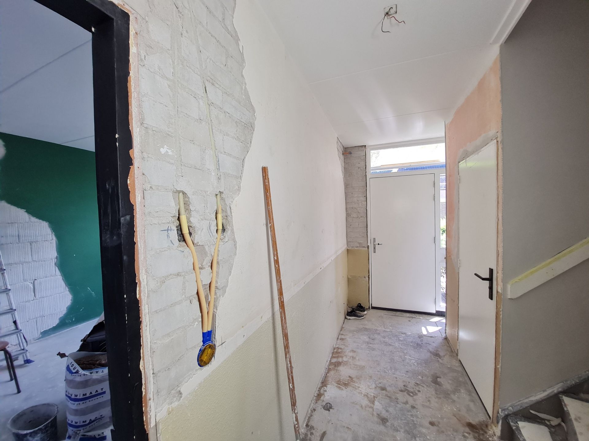 Smalle gang in renovatie met blootliggende bedrading, afgebladderde muren, puin op de vloer en een deur aan het einde.
