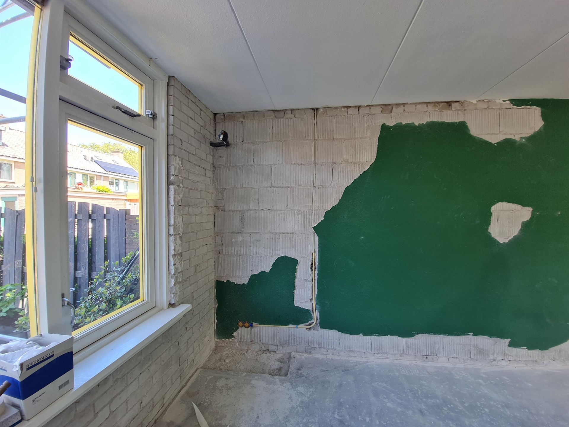 Een gedeeltelijk ontdekte kamermuur met groene verf, zichtbaar wit stucwerk en een raam aan de linkerkant.