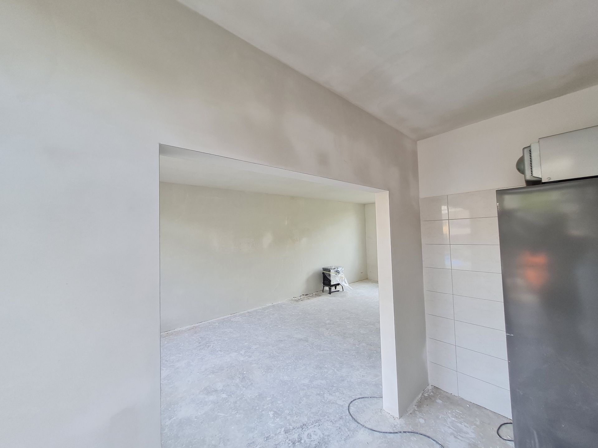 Een lege, witte kamer met een brede deuropening, een lichte vloer en een donker apparaat aan de rechterkant.