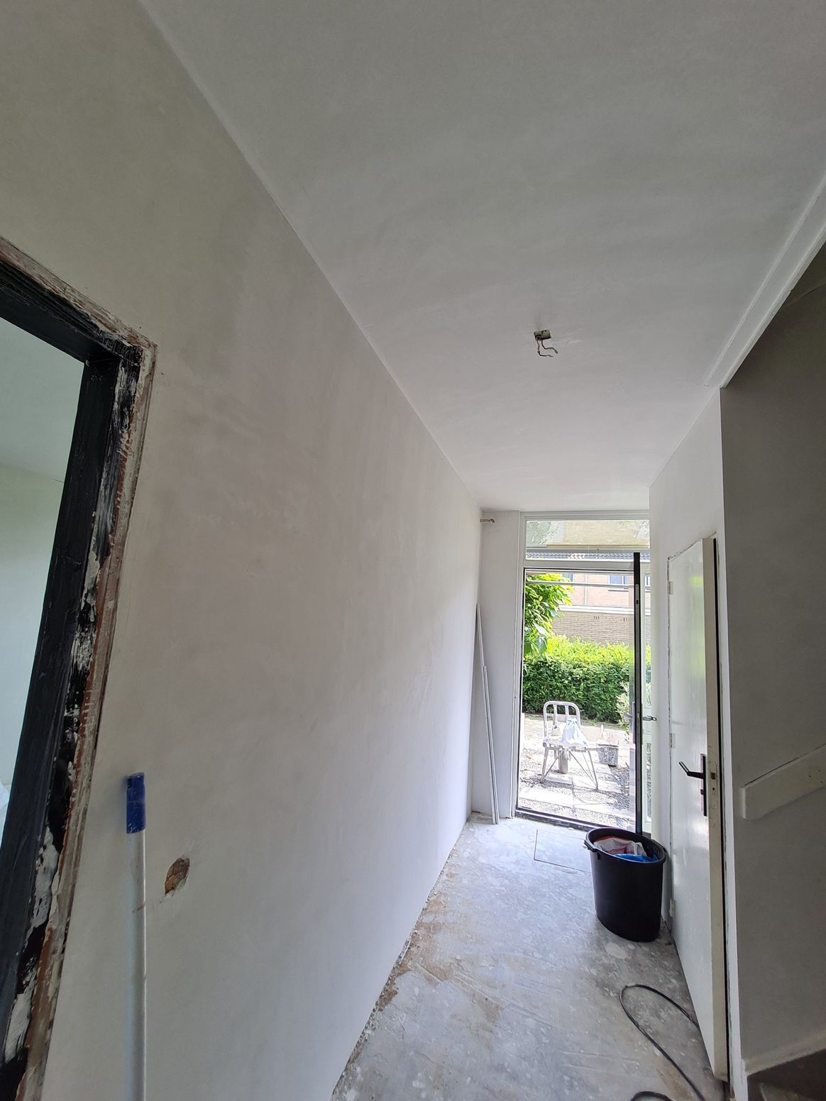 Smalle gang in renovatie met witte muren, onafgewerkte vloer en een open deur naar een lichte tuin.