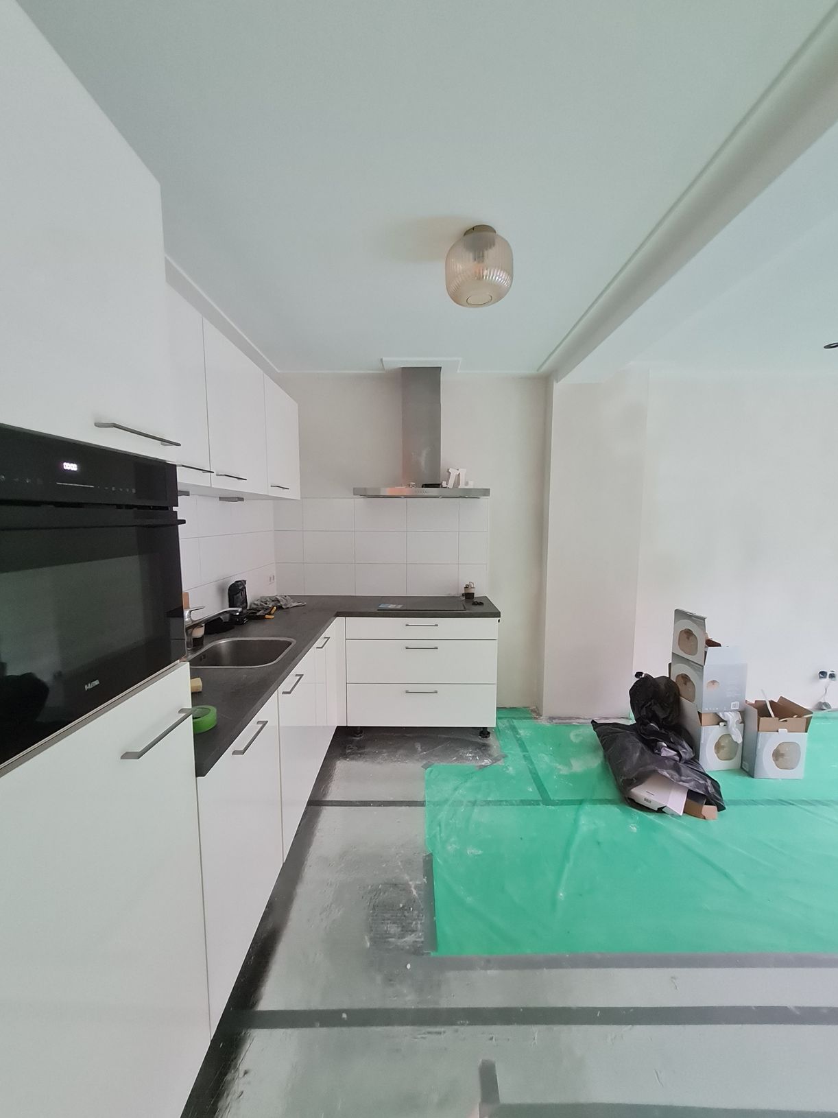 Een lichte, witte keuken met zwarte apparaten en een groen zeil op de vloer, vlakbij gereedschap en tassen.