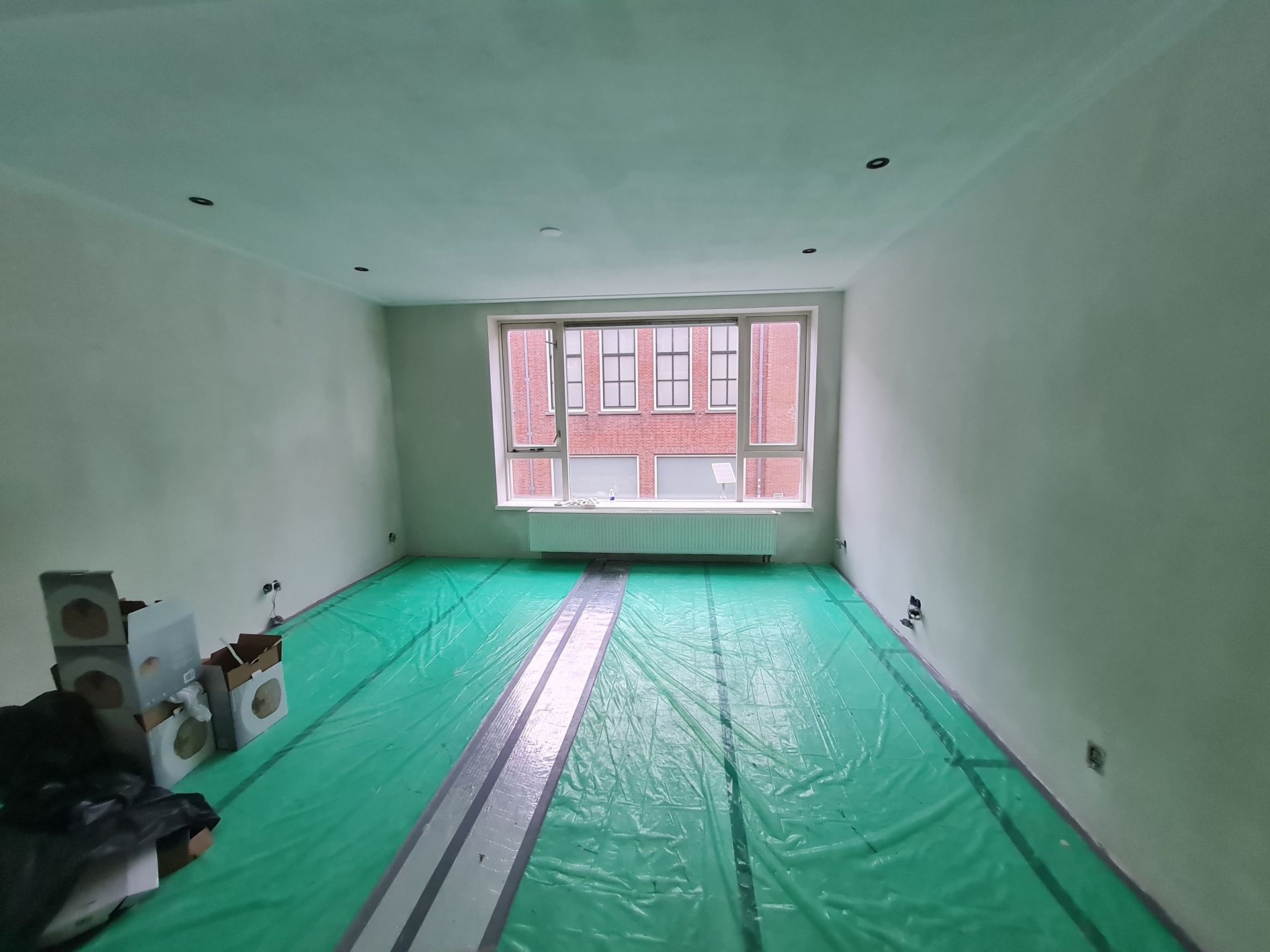 Een lege kamer die gerenoveerd wordt, met een groene vloerbedekking, witte muren en een raam aan de achterkant.