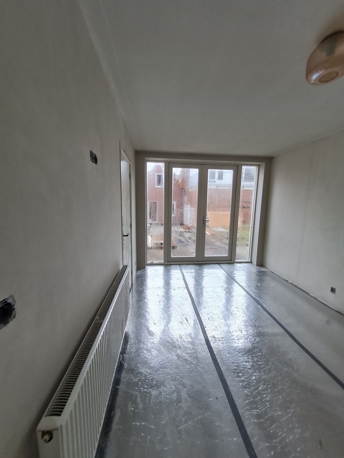 Lege, smalle kamer met witte muren, radiator en grote glazen deuren die toegang geven tot een balkon.