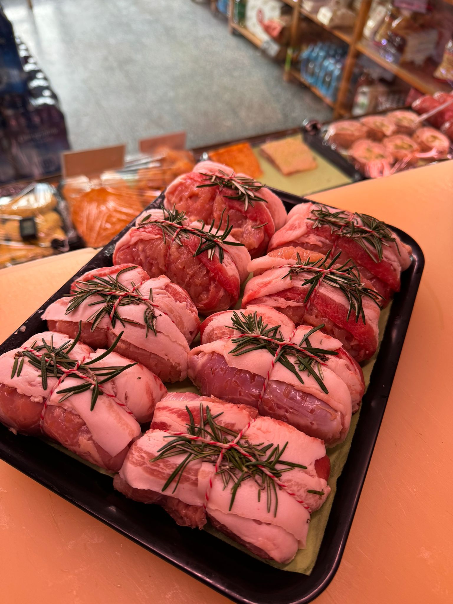 Vassoio di carne cruda, avvolta nella pancetta e guarnita con rametti di rosmarino, in un ambiente da gastronomia.