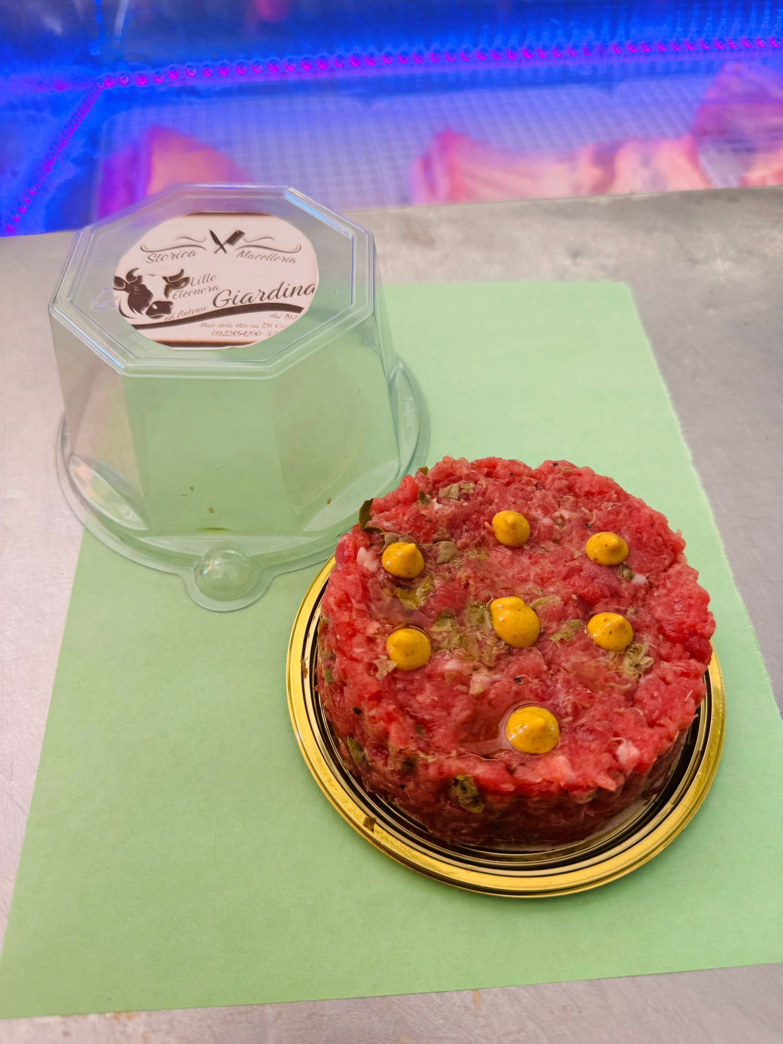 Tartare di carne cruda decorata con puntini gialli, accanto a un contenitore di plastica trasparente, su una superficie verde.