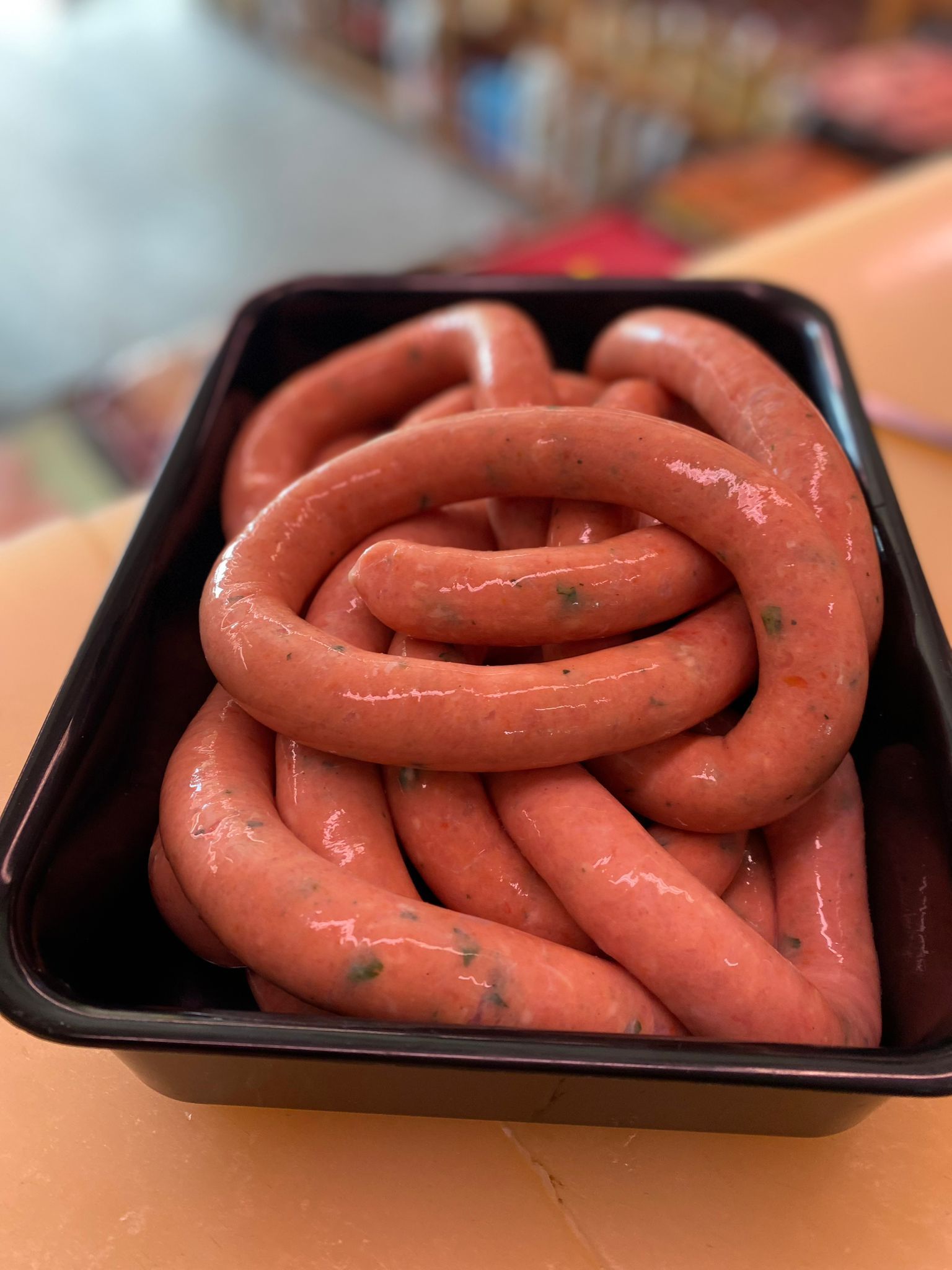 Salsicce crude in un contenitore di plastica nera, strettamente arrotolate. Le salsicce sono rosa con erbe aromatiche visibili, disposte su una superficie di colore chiaro.