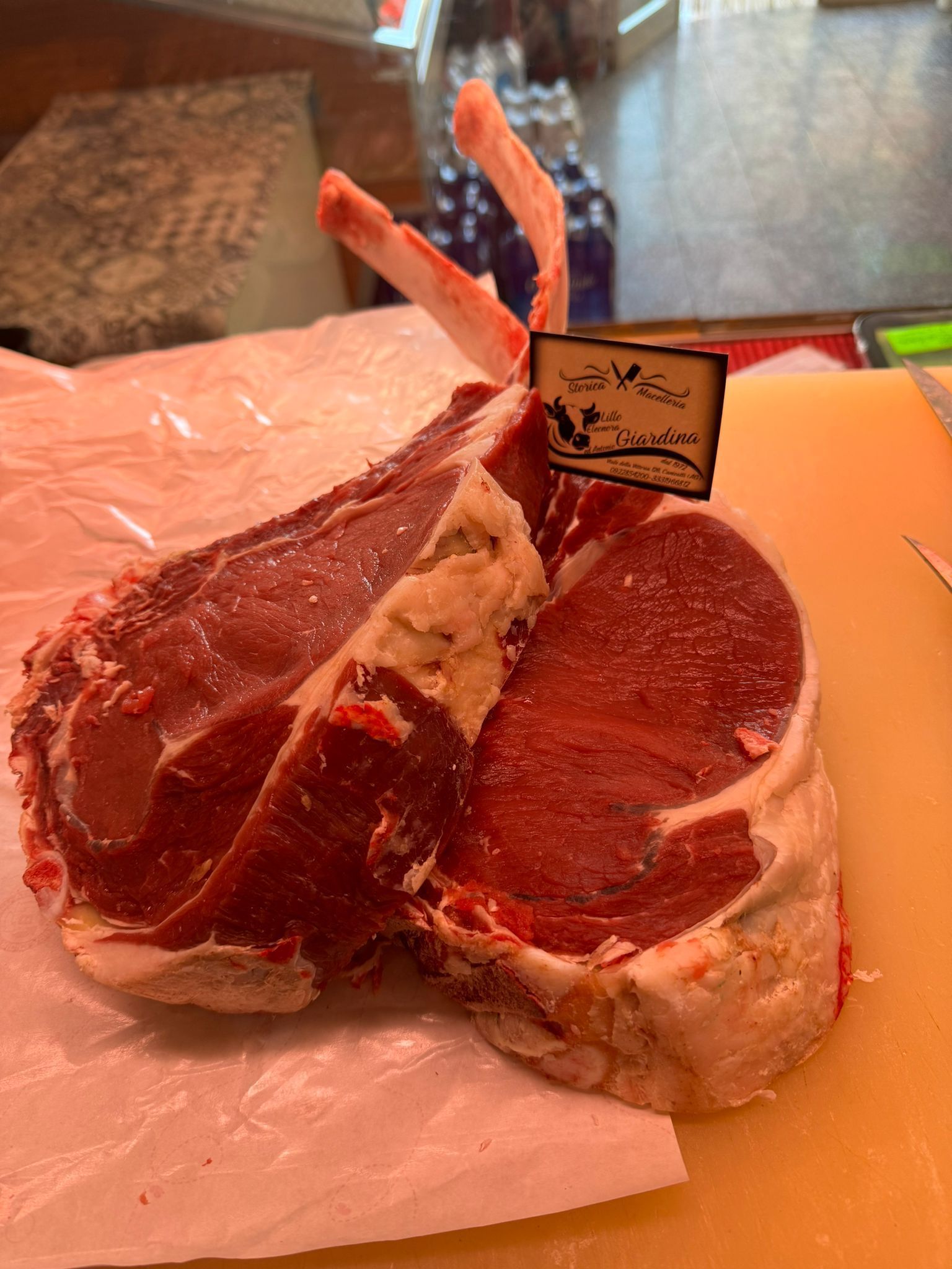 Bistecca di ribeye con osso e etichetta, appoggiata su carta da macellaio e su un tagliere giallo.