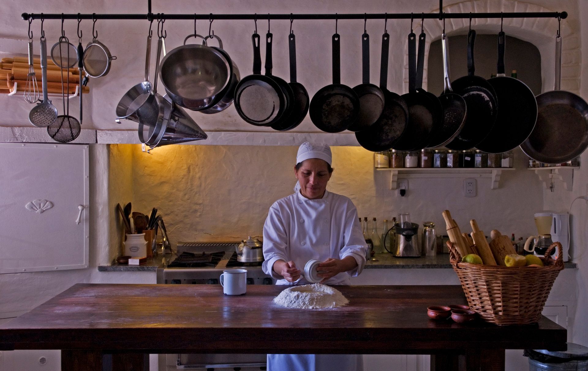 A chef in a white uniform prepares dough on a wooden table in a kitchen with hanging pots and a basket of bread.