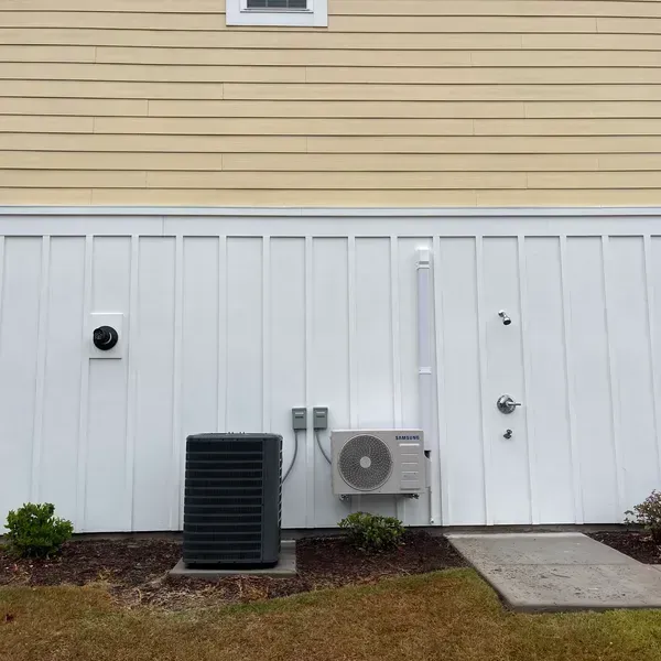 White house siding with utility units and side door beside a small concrete pad and patchy lawn.