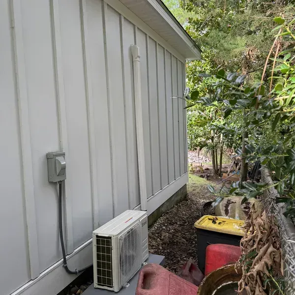 Side of a white house with an outdoor air conditioner unit beside a red trash bin and trees.