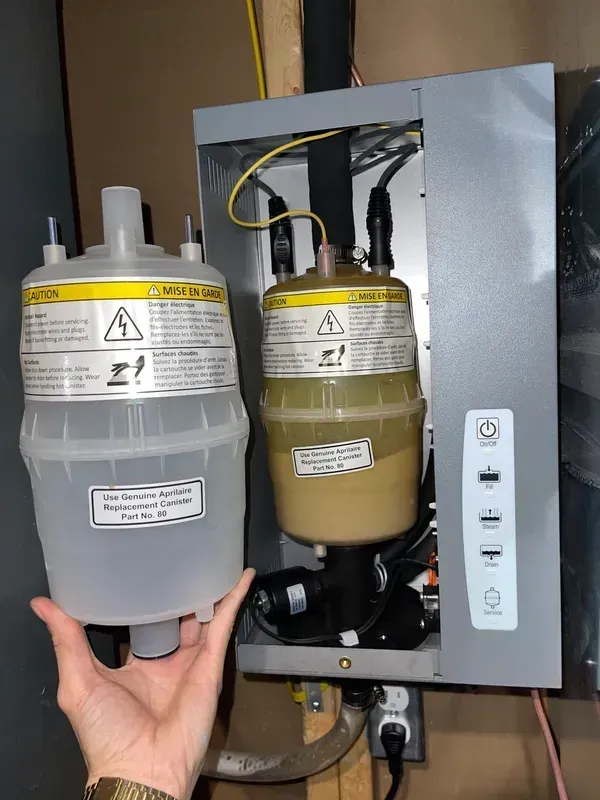 Hand holding a gray container beside a tan filter canister inside an open cabinet