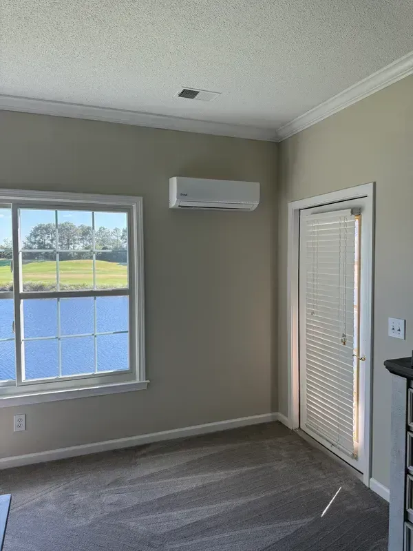 Empty beige room with window, wall-mounted AC, and blinds-covered door with carpeted floor