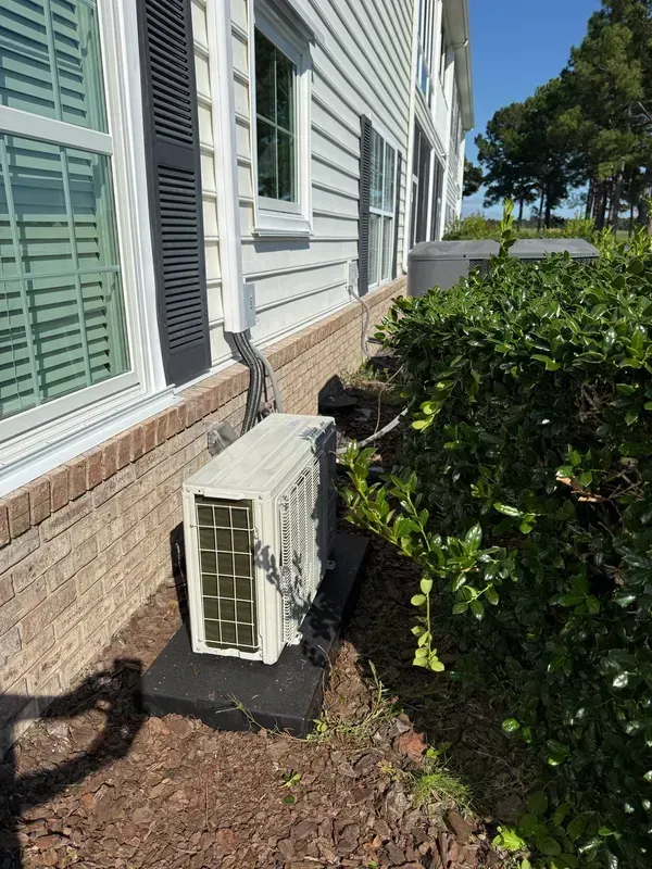 Outdoor HVAC unit beside a house wall with green shutters and nearby bushes