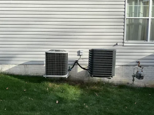 Two outdoor air conditioning units beside a house wall and window, with grass in front.
