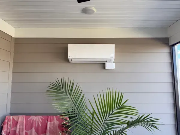 Outdoor patio with wall-mounted air conditioner, potted palm, and covered furniture against beige siding