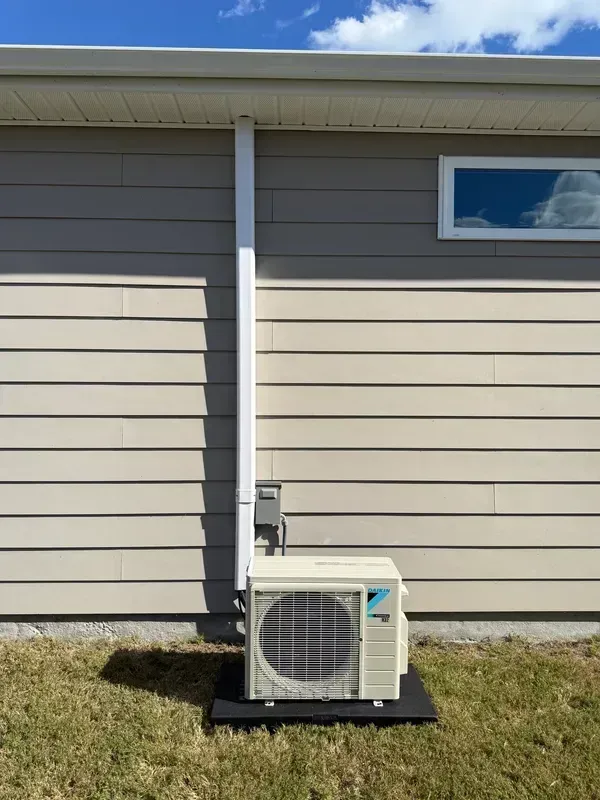 House exterior with a white utility unit on a black pad beneath a downspout, beside beige siding and a small window