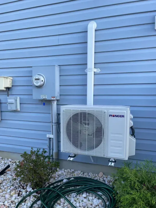 Blue house exterior with a mounted mini-split AC unit, utility meter, and coiled green hose near rocks.