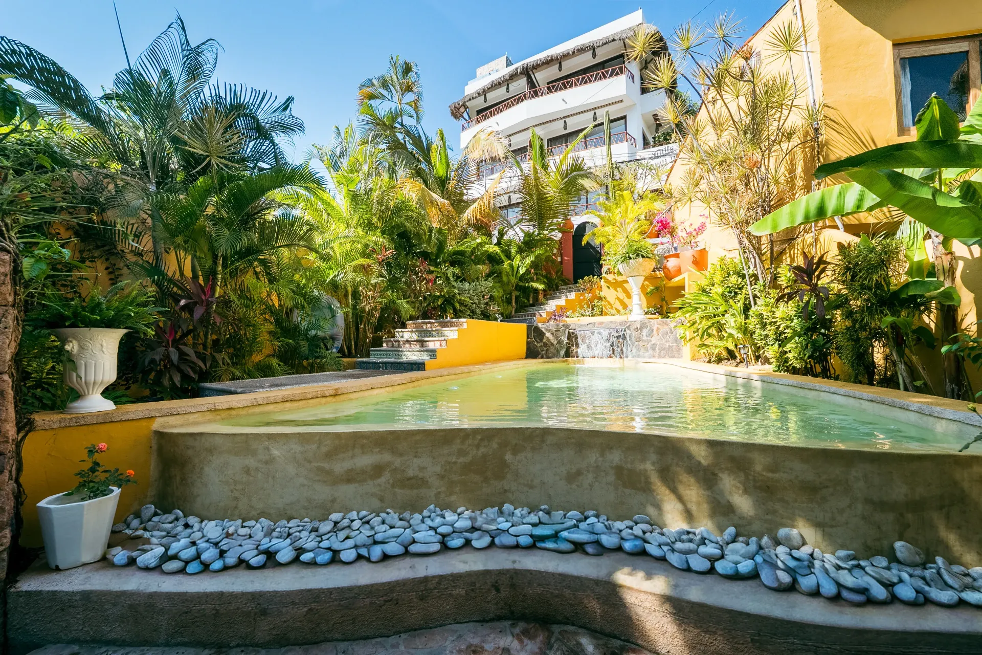Swimming pool in a tropical garden with cascading water feature, white building in the background.