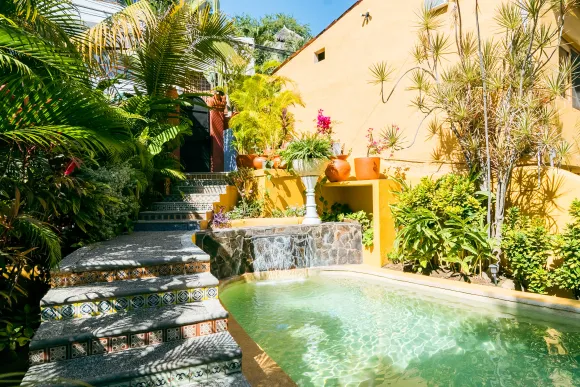 Stone steps lead up to a yellow building with potted plants and a pool with a small waterfall.