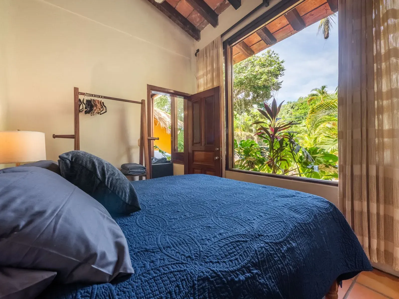 Bedroom with a blue bed, open door, and large window overlooking lush greenery.