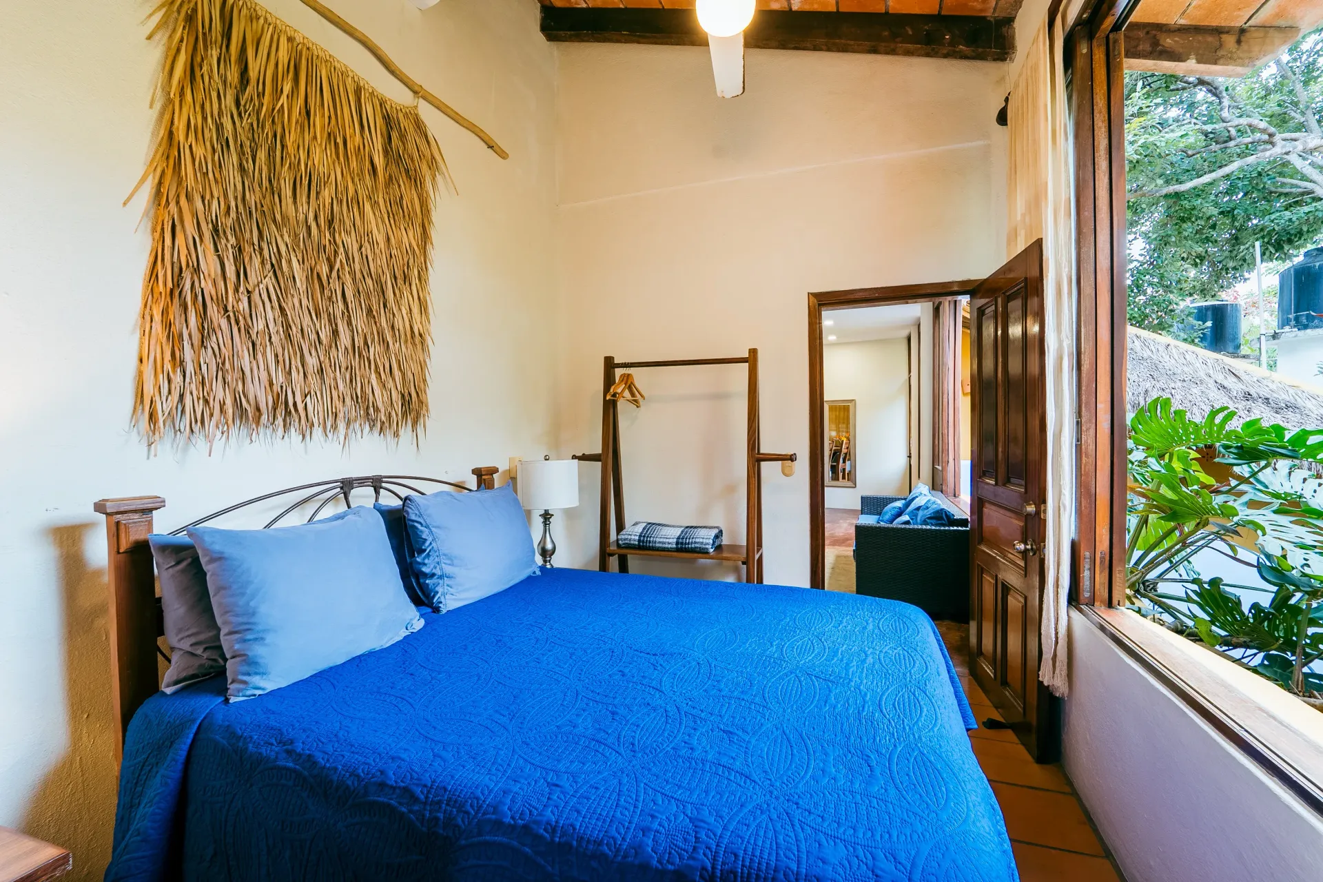 Bedroom with blue bed, natural wall art, open door to another room, and outdoor view.