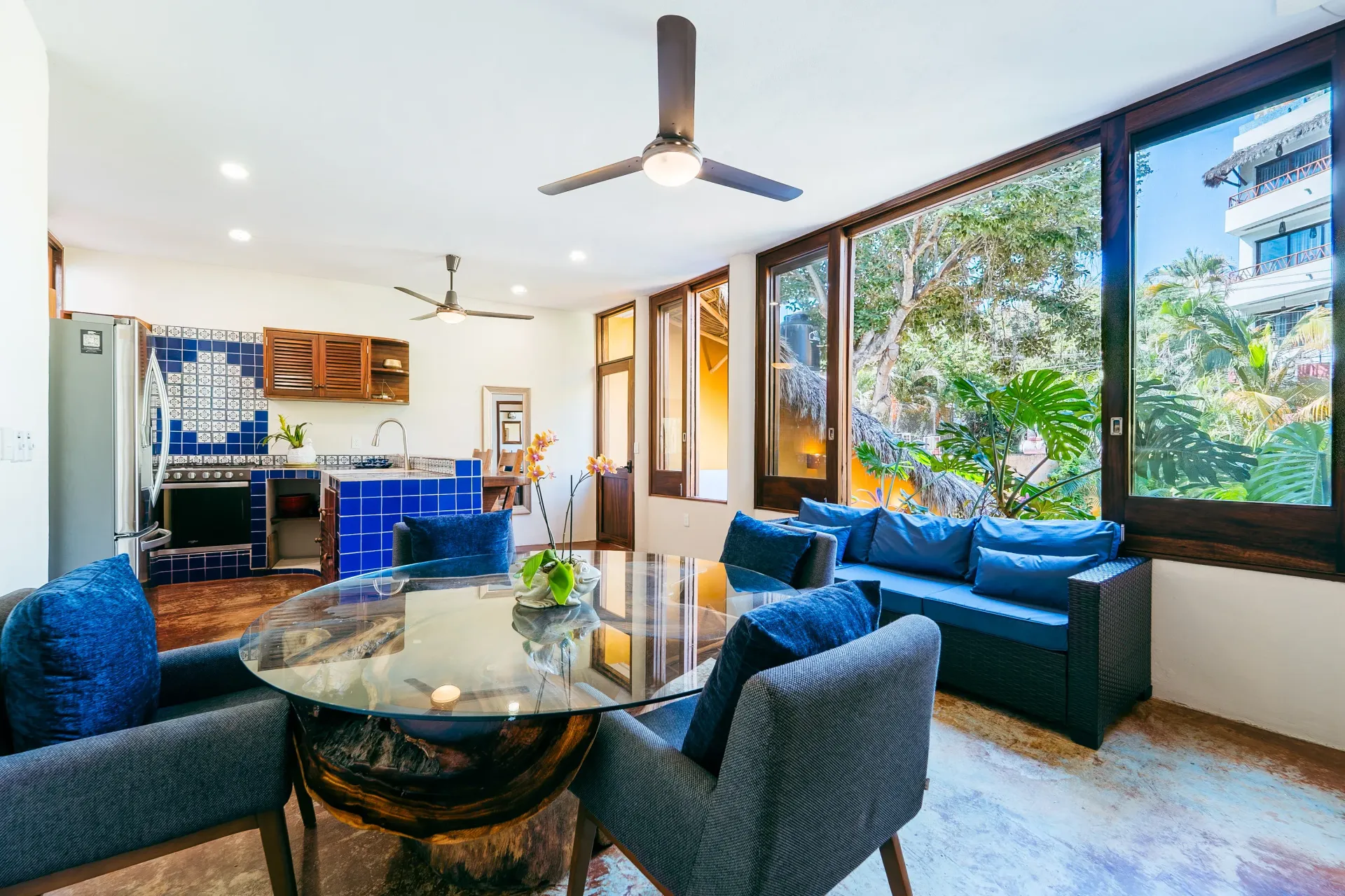 Living area with dining table, blue chairs, sofa, and a kitchen. Large windows overlook greenery.