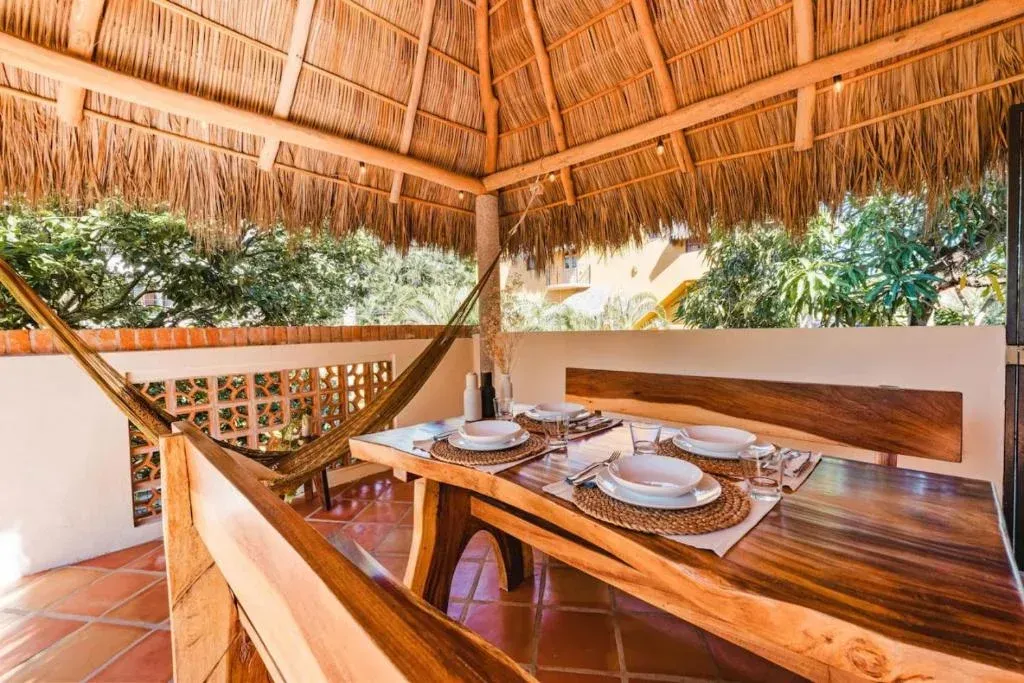 Outdoor dining area with a thatched roof, wooden table and benches, and a hammock.