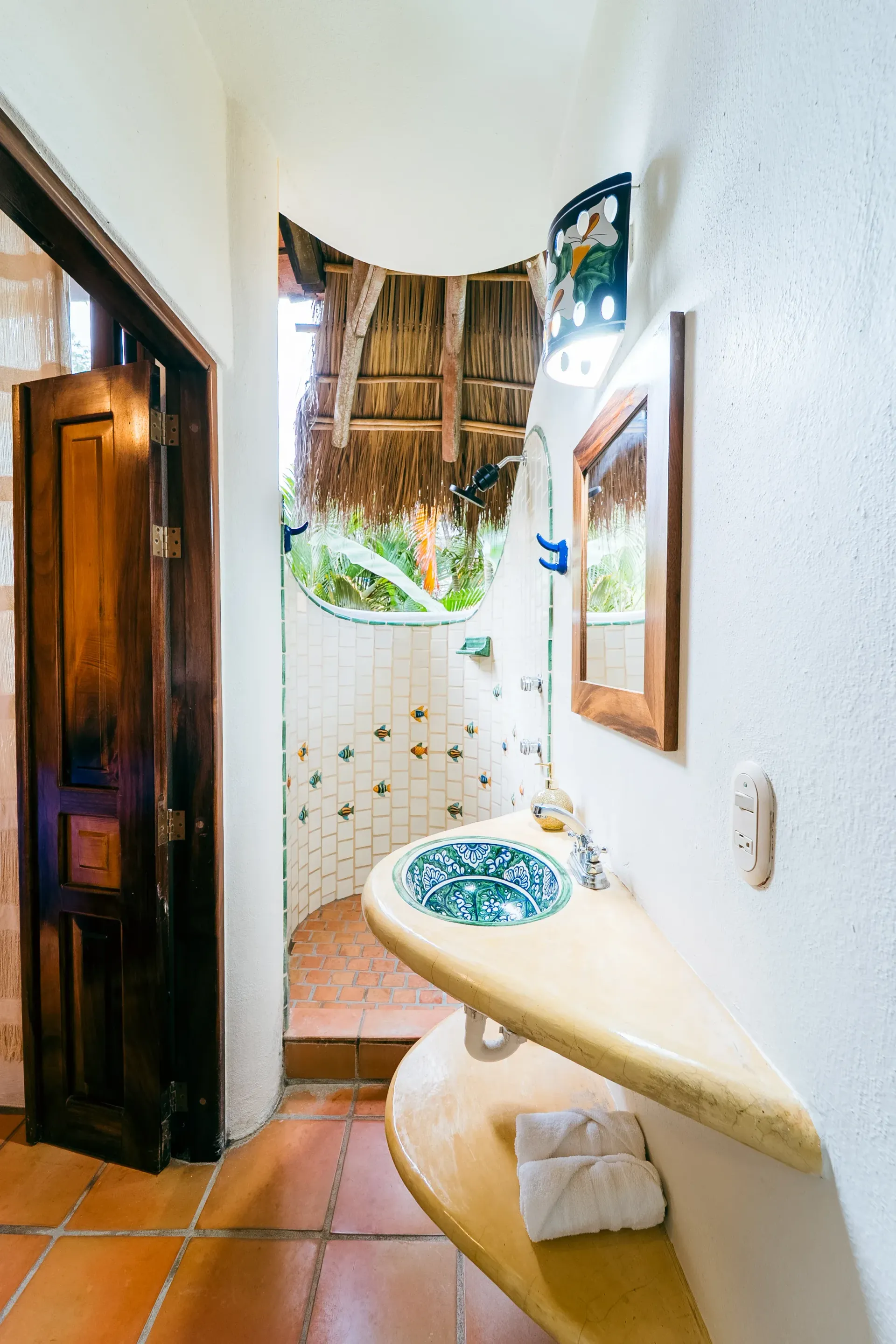 Bathroom with curved countertop, patterned sink, thatched roof shower opening, wood door, terracotta floor.