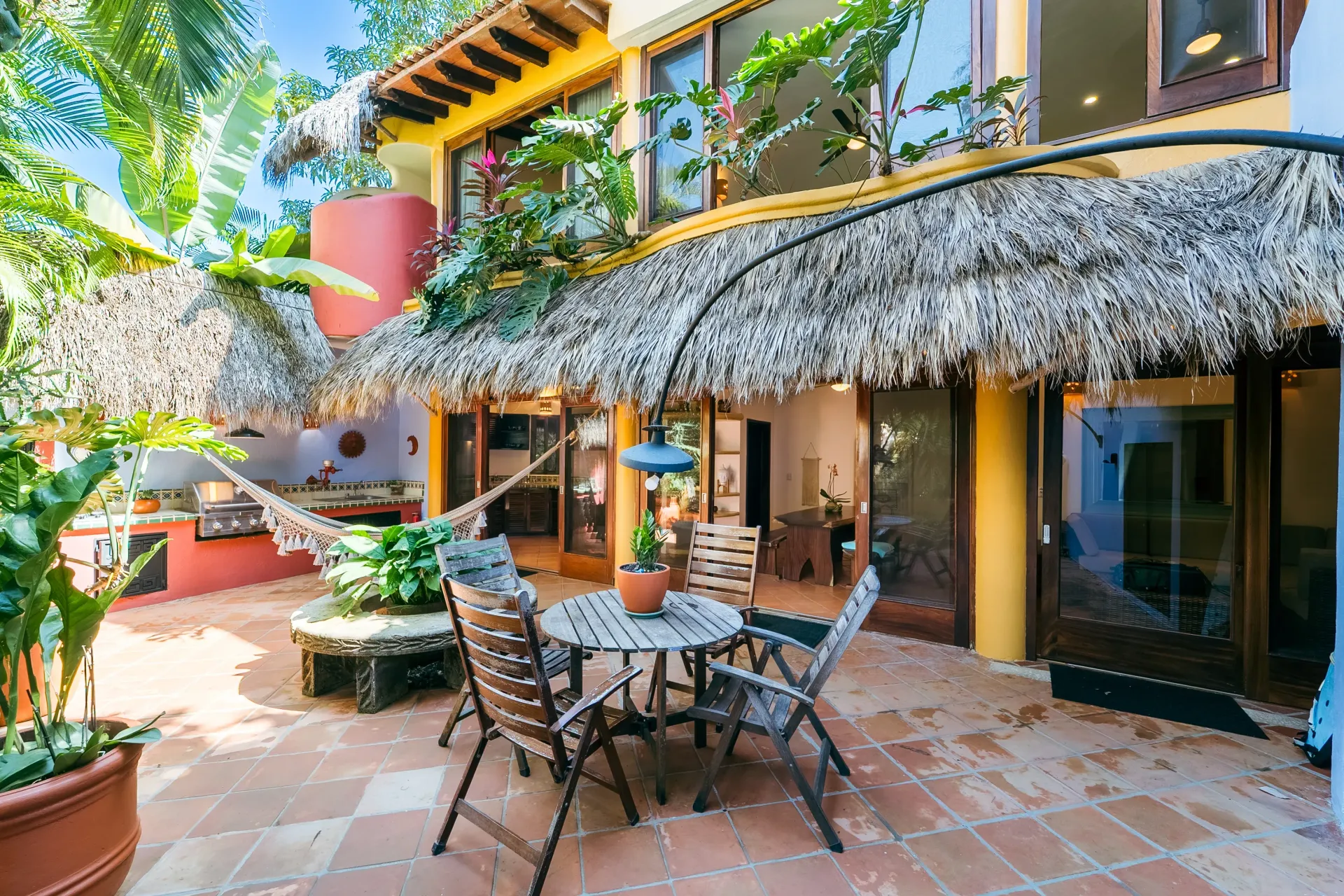 Patio with thatched roof, table and chairs, hammock, and building with glass doors and windows.