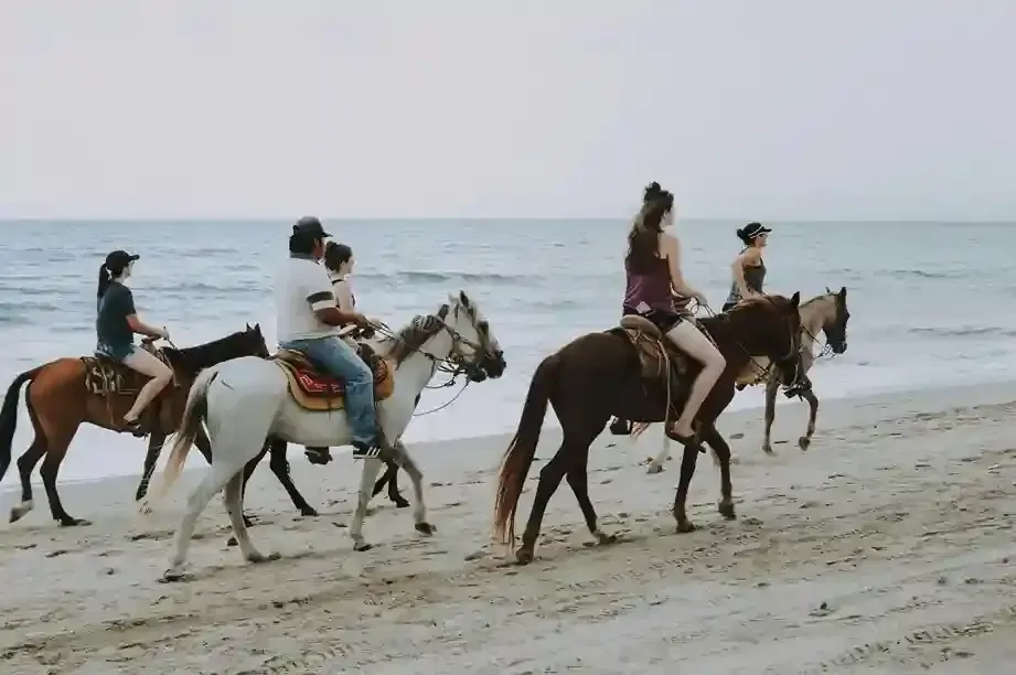 People horseback riding on a sandy beach near the ocean; overcast sky.