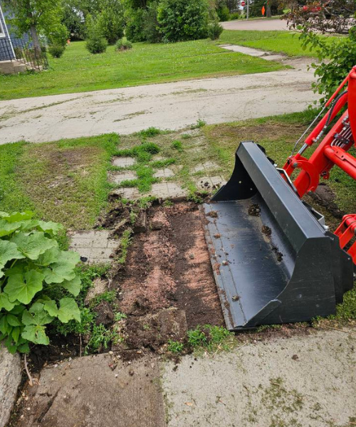 A tractor bucket digging in a dirt area beside a concrete path and grassy yard.