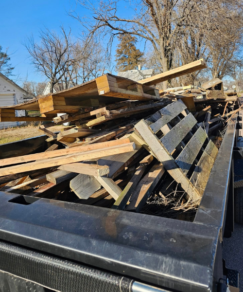 Pile of wooden planks and pallets in the bed of a black pickup truck, outdoors under a sunny sky.