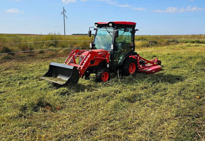 Red tractor with a front-end loader and mower cutting grass in a field on a sunny day.