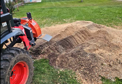 A tractor with an orange stump grinder is removing wood from a stump, creating a large pile of wood chips on a grassy lawn.