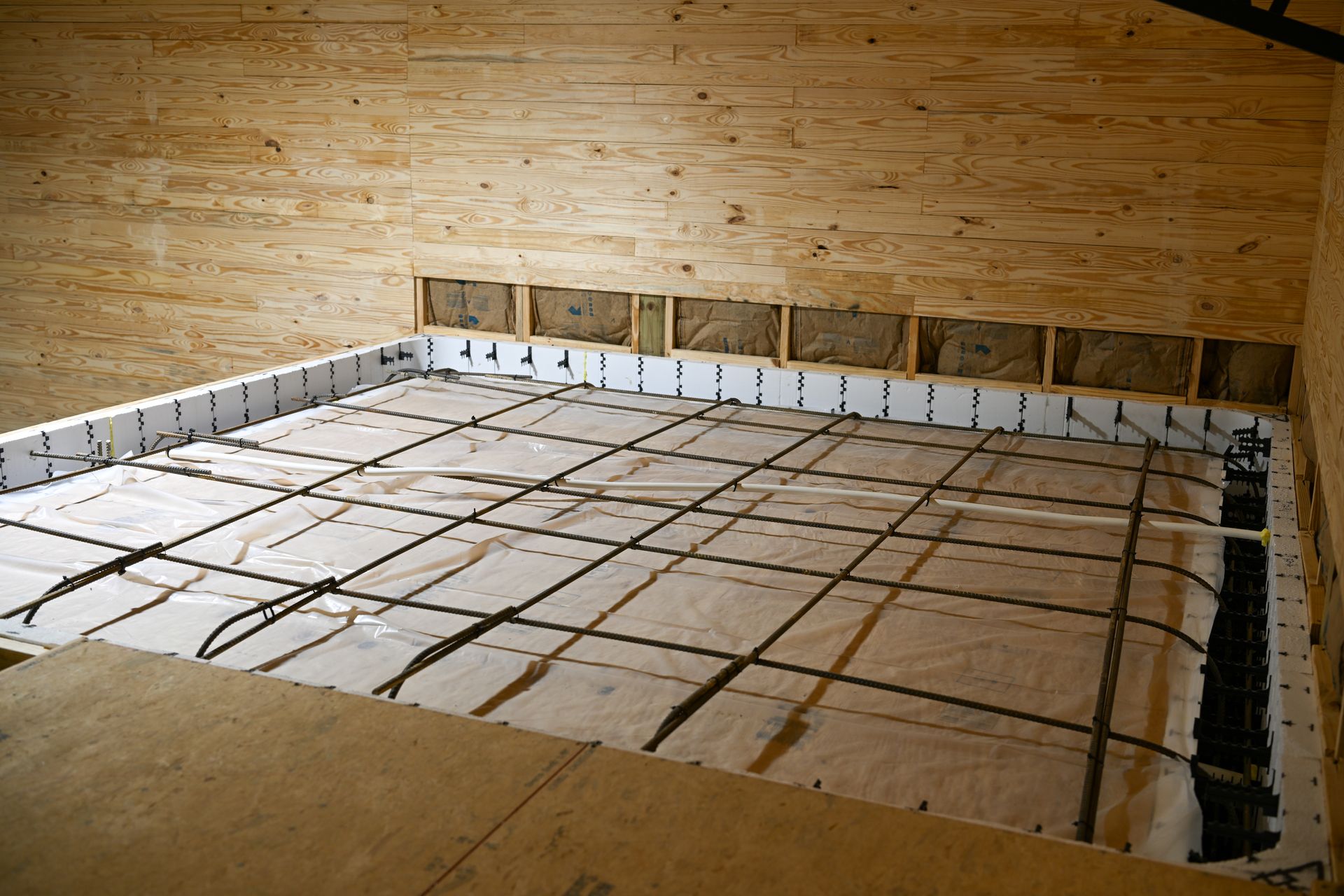 A view of a concrete floor foundation being prepared with a rebar grid, white vapor barrier, and foam insulation boards. A view of a concrete floor foundation being prepared with a rebar grid, white vapor barrier, and foam insulation boards.