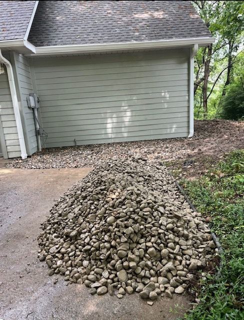 A pile of gray river rocks on a wet paved driveway in front of a green-sided building.