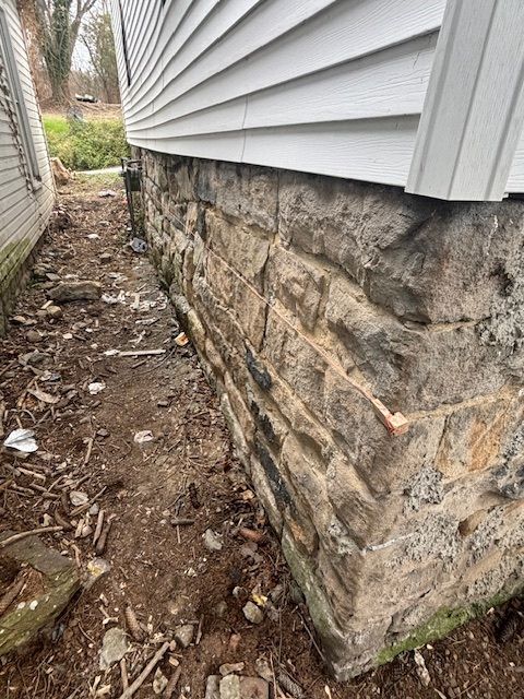 Stone foundation of a house with white siding and a narrow dirt yard.