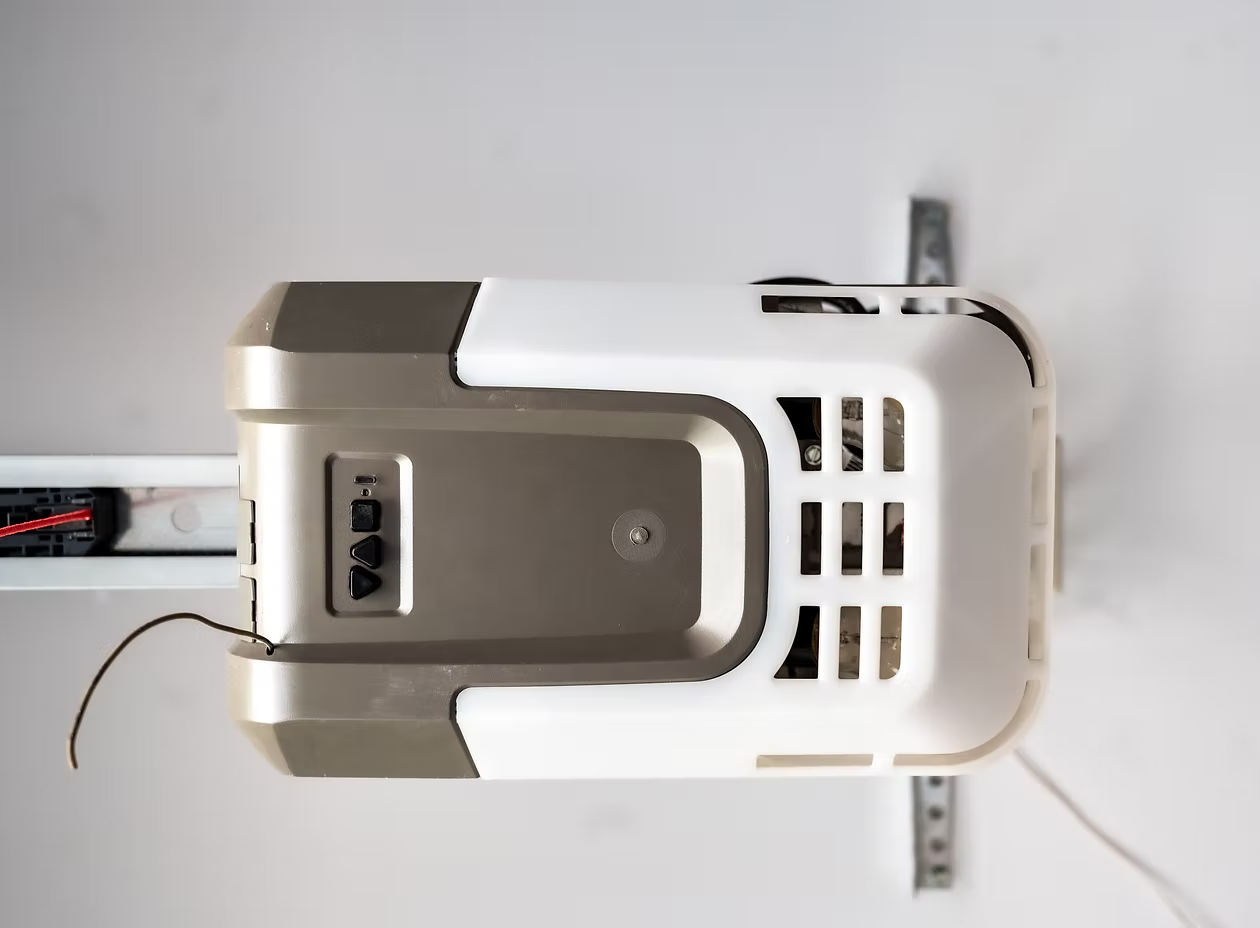 A garage door opener motor unit mounted to the ceiling, featuring a white and silver housing with a hanging antenna wire.