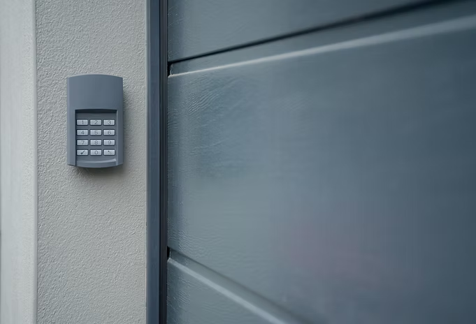A gray keypad mounted on a light-colored exterior wall next to a modern gray garage door.