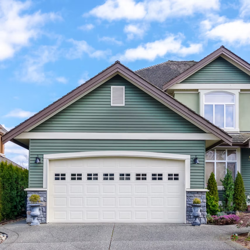 A suburban house with green siding, a large white two-car garage door, and a concrete driveway under a blue sky.