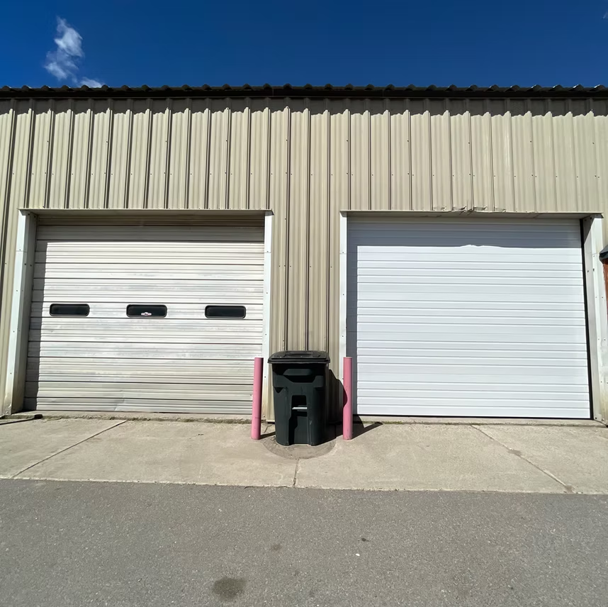 A beige metal building with two white roll-up garage doors separated by a black trash bin and two pink bollards.