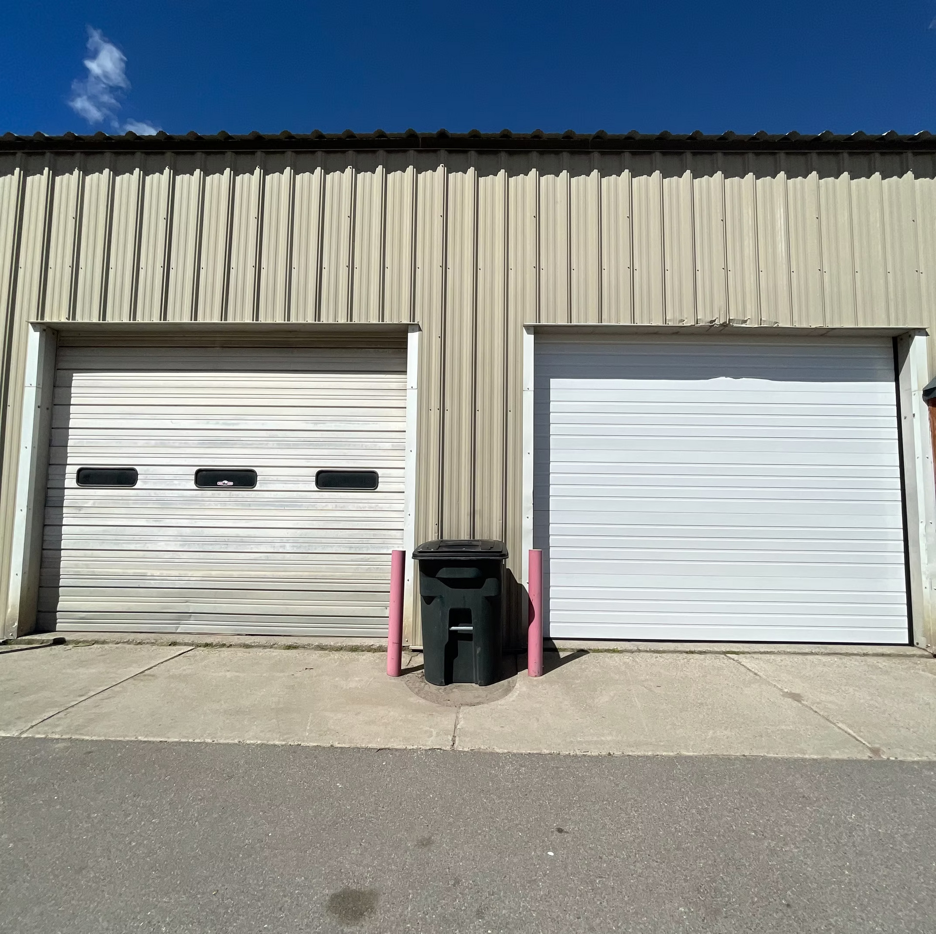 Two closed white garage doors on a beige corrugated metal building with a single black trash bin between them.