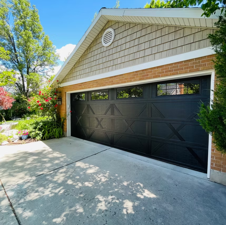 A black carriage-style garage door with windows on a brick and shingle house exterior, framed by green trees and bushes.